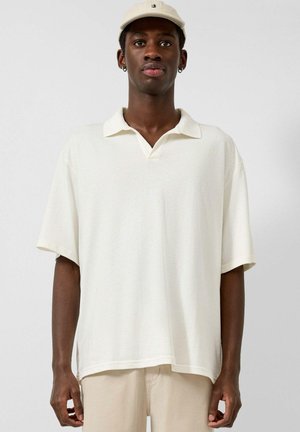 Młody mężczyzna w kremowej koszulce polo z krótkim rękawem, beżowych spodniach i beżowej czapce z daszkiem, stoi na jednolitym tle.