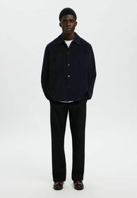 Hombre de pie con las manos en los bolsillos, vistiendo una chaqueta abotonada de color azul marino oscuro, pantalones negros, camiseta blanca y zapatos de cuero oscuros sobre un fondo blanco.
