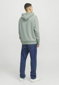 Felpa grigia con cappuccio dal taglio rilassato, abbinata a jeans in denim blu e sneaker bianche, vista da dietro.