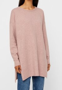 Frau trägt einen lockeren, langärmligen, hellrosa Pullover mit seitlichen Schlitzen über blauen Jeans und steht vor einem schlichten weißen Hintergrund.