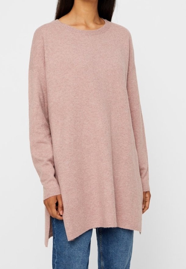Frau trägt einen lockeren, langärmligen, hellrosa Pullover mit seitlichen Schlitzen über blauen Jeans und steht vor einem schlichten weißen Hintergrund.