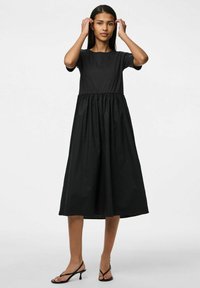 Crna haljina s uskim tijelom i naboranom, lepršavom suknjom. Kratki rukavi, okrugli izrez, izrađena od mekog materijala. Nosena uz crne sandale na remen.