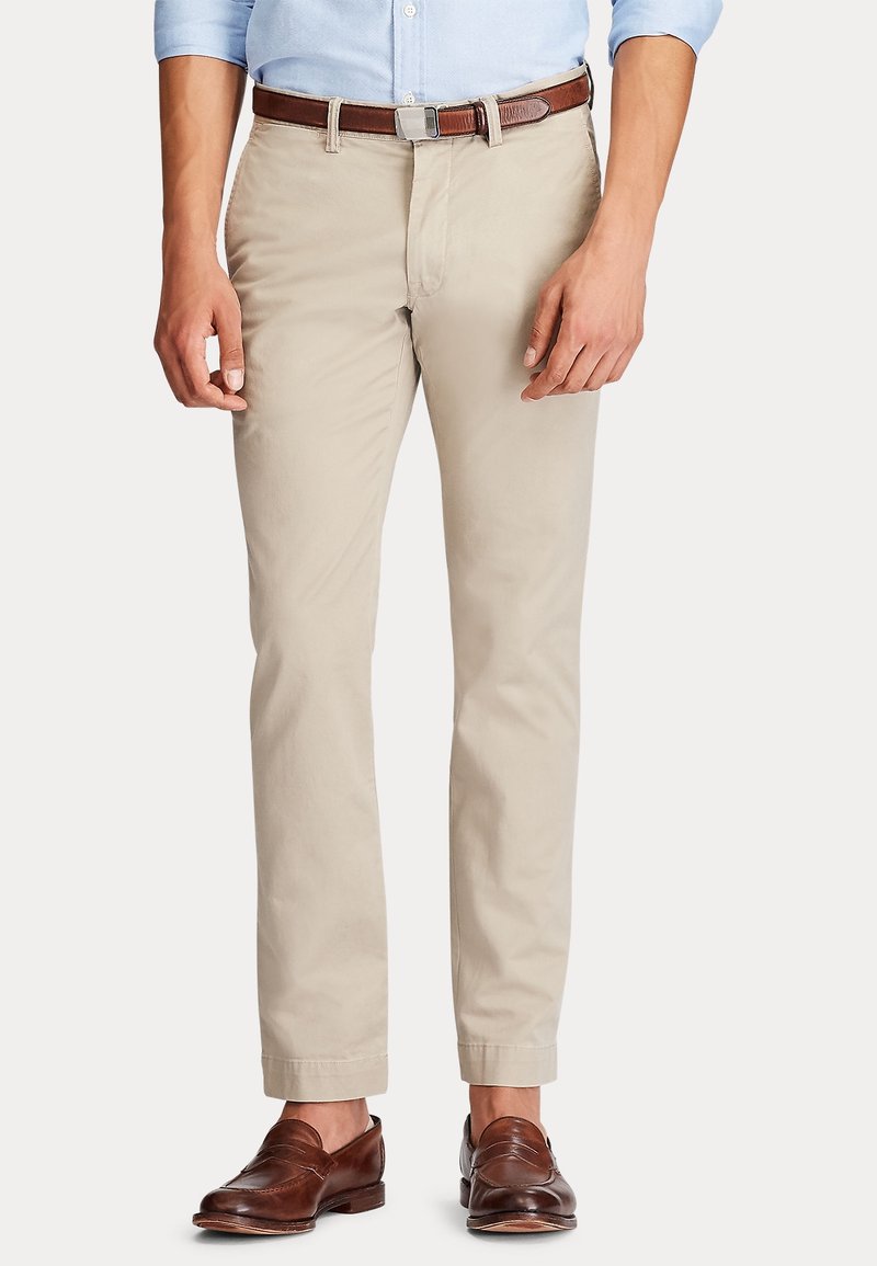 Homme portant un chino beige, une ceinture en cuir marron et des mocassins en cuir marron, debout, les mains détendues le long du corps.