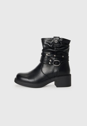 Bottine en cuir noir avec un haut replié, une sangle décorative avec des accents métalliques, un bout rond et un large talon. Texture lisse.