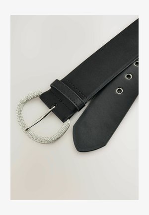 Mustvalge nahkrihm, millel on kaarduv hõbeplaat, kaunistatud sädelevate kivikestega. Rihmal on sile tekstuur ja mitmed augud reguleerimiseks.