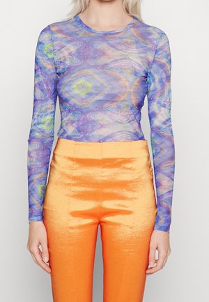 Kvinde iført langærmet, flerfarvet mønstret top med orange højttaljede, skinnende tætsiddende bukser, der står med arme afslappede.