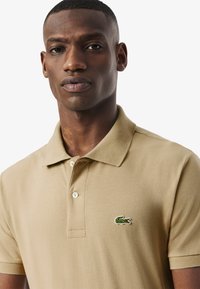 Beige polo skjorta gjord av texturerad tyg. Har en vikbar krage och tre knappar, med en liten grön broderad krokodil-logotyp på bröstet.