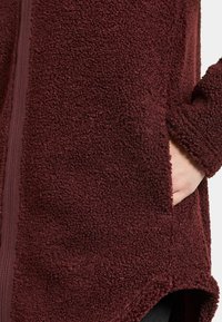 Burgundy fleecejacka med en mjuk, texturerad yta, dragkedjestängning och sidofickor med en asymmetrisk fåll.