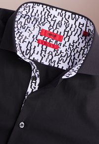 Schwarzes Hemd mit Klappkragen, das das schwarz-weiß gemusterte Innenfutter und rote "HUGO" Markenetiketten zeigt.