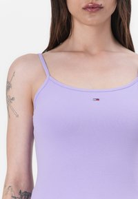 Lila toppi ohuilla spagettiolkain, valmistettu sileästä materiaalista. Rinnassa on pieni logo, jossa on punaisia, valkoisia ja sinisiä värejä.