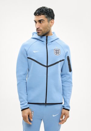 Homme portant un survêtement sportif bleu clair de l'Angleterre avec capuche, fermeture éclair noire et logos, regardant de côté sur fond blanc.