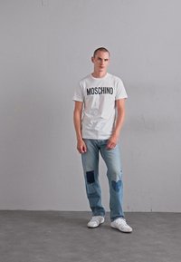 Mees seisab halli seina äärde toetunult, seljas valge Moschino T-särk, helesinised teksad tumepruunide laikudega ja valged tennisetossud betoonpõrandal.
