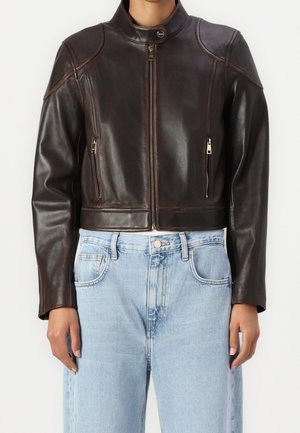 Giacca di pelle marrone scuro con cerniera frontale e tasche con cerniera indossata sopra jeans azzurri a vita alta su una persona in piedi.