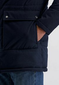 Marineblauwe pufferjack met een grote voorklep met drukknoopsluiting, zachte textuur en geborduurde ontwerpen. Gedragen over denim blauwe jeans.