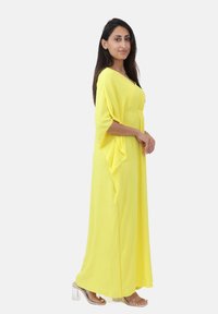 Robe maxi jaune en tissu léger avec un design fluide, des manches courtes et une taille cintrée. Associée à des sandales à talons transparents.