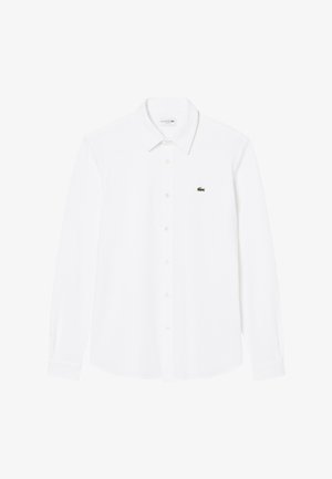 Balts garo piedurknēm kreklis, izgatavots no kokvilnas. Izceļas ar klasisku jaku un mazu zaļu Lacoste logotipu uz kreisā krūšu puse.