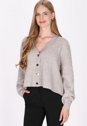Lichtgrijze geribde cardigan met een V-hals, oversized pasvorm en vijf metalen knopen aan de voorkant, gecombineerd met zwarte broeken.