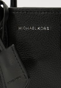 Černá kožená taška s texturou, která má vyražené logo "MICHAEL KORS" ve stříbrné barvě, s hladkým koženým štítkem připevněným k ní.