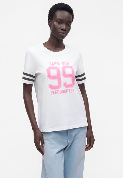 Γυναίκα με λευκό T-shirt με ροζ «99» και κείμενο «BAUM UND PFERDGARTEN», σε συνδυασμό με ανοιχτό μπλε τζιν, στέκεται μπροστά σε απλό λευκό φόντο.