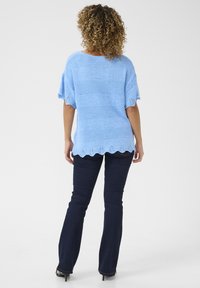 Modra pletena majica s kratkimi rokavi in zapletenim valovitim robom, v kombinaciji s temnimi kavbojkami. Tkanina ima teksturiran videz.