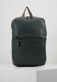 Sac à dos en cuir noir avec poche zippée à l'avant et surface texturée, se tenant debout sur un piédestal blanc contre un fond clair.