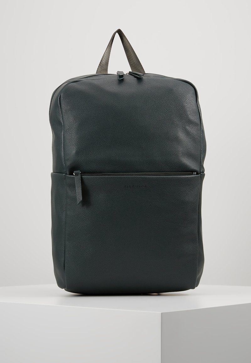 Sac à dos en cuir noir avec poche zippée à l'avant et surface texturée, se tenant debout sur un piédestal blanc contre un fond clair.