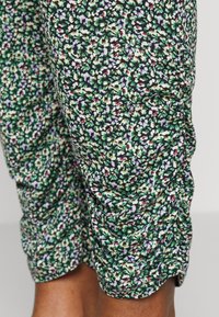 Calças capri florais com um padrão verde, preto e branco, apresentando um tecido texturizado e detalhes franzidos nas laterais.