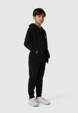 Ragazzo in piedi con le mani nelle tasche, indossa una felpa nera con zip, pantaloni neri e sneakers bianche con velcro, su uno sfondo chiaro e uniforme.