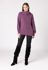 Maglione spesso di colore viola con colletto a scialle, maniche lunghe, orlo a costine, indossato con jeans skinny neri e stivali neri alti sbuffanti.