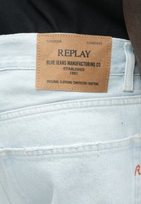 Lyseblå denimjeans med et lærmerke som viser "REPLAY" og detaljer om merket. Iøynefallende sømmer og lommer synlige.