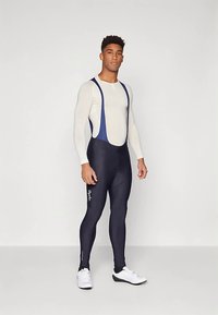 Giovane in piedi, indossando una camicia bianca a maniche lunghe, pantaloni da ciclismo blu scuro con logo del marchio e scarpe da ciclismo bianche su sfondo neutro.