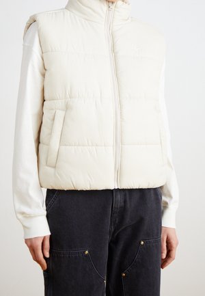 Personne portant une veste matelassée blanche sans manches sur un haut blanc à manches longues et un pantalon noir avec poches avant et détails dorés.