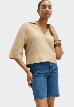 Kvinde iført beige V-hals, top med korte ærmer og blå denim knælange shorts, accessoriseret med armbånd af træ og perler.