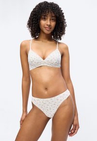 Γυναίκα με σγουρά μαλλιά που φοράει λευκό σουτιέν με λουλουδάτο σχέδιο και ταιριαστό εσώρουχο, σε απλό φωτεινό φόντο.