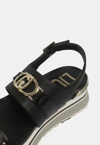 Sandalo in pelle nera con cinturino regolabile, dettagli in hardware dorato, accenti texture e una suola piatta bianca con un motivo sottile.