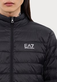 Veste matelassée noire avec un col haut, fermeture éclair et design couturé. Présente un logo EA7 Emporio Armani blanc sur la poitrine.