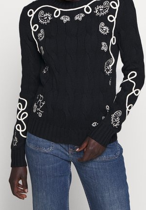 Pull en maille torsadée noir avec broderies blanches paisley et boucles, porté avec un jean bleu sur une personne à la peau foncée.