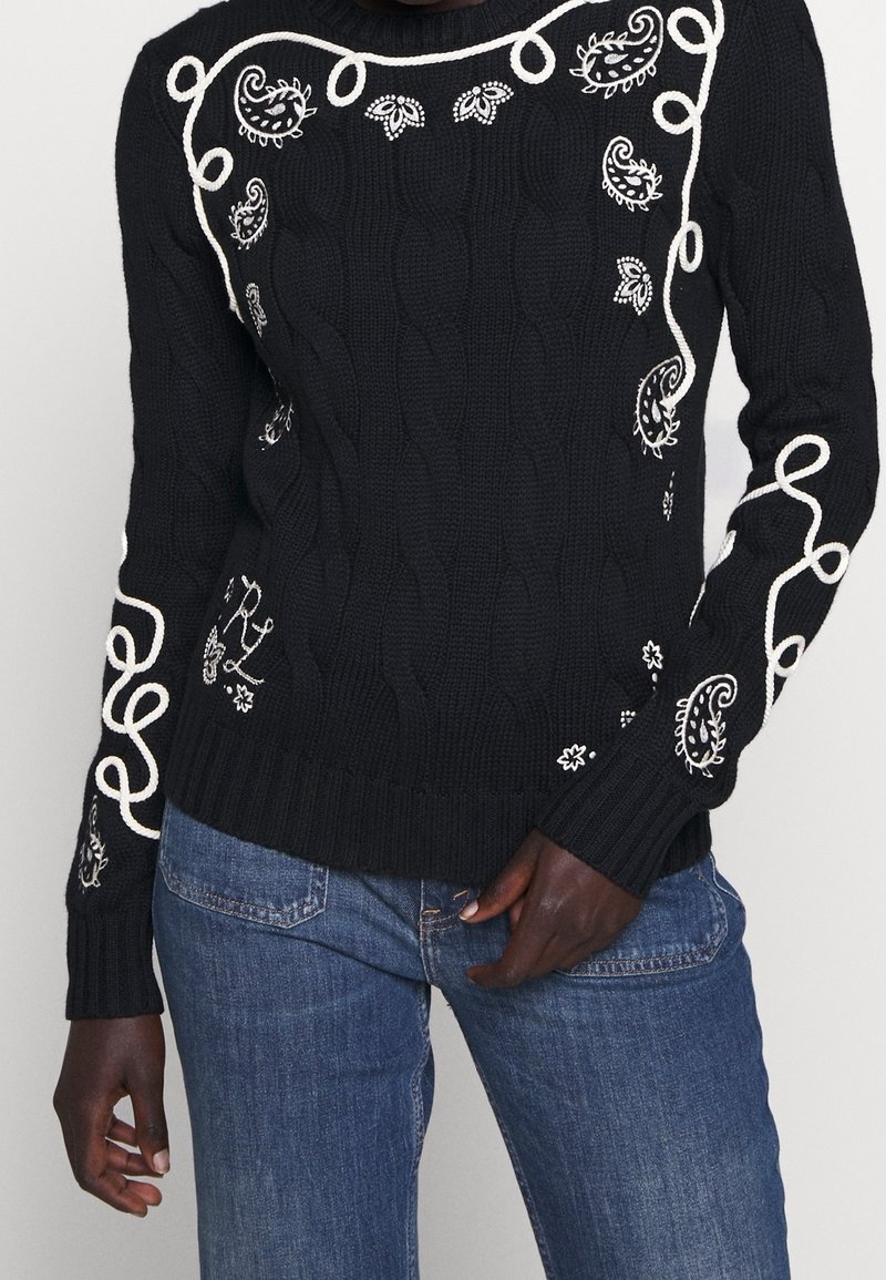 Pull en maille torsadée noir avec broderies blanches paisley et boucles, porté avec un jean bleu sur une personne à la peau foncée.