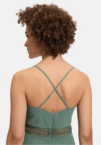 Zielona sukienka z regulowanymi krzyżującymi się ramiączkami, koronkowym detalem z siateczki w talii oraz ukrytym zapięciem z tyłu.