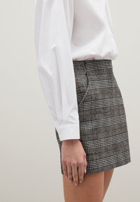 Grau karierte Minirock mit braunen Akzenten, ausgestattet mit Seitentaschen und einer strukturierten Passform. Kombiniert mit einem weißen Langarmshirt.