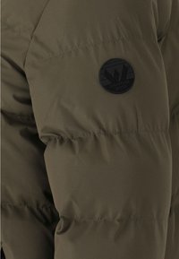 Chaqueta de plumas verde oliva con textura acolchada. Presenta un parche de logo negro redondo con un diseño geométrico en la parte superior de la manga izquierda.