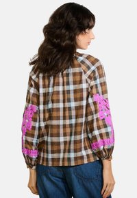 Camicia a quadri marrone e bianca con ricami floreali rosa sulle maniche. Vestibilità ampia, maniche lunghe e dettagli arricciati ai polsi. Design casual.