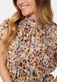Blumige Rüschbluse in Beige mit braunen und blauen Punkten. Leichtes Material mit Knopfverschluss und gefalteten Akzenten am Ausschnitt.