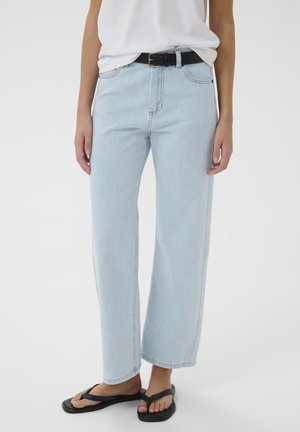 Personne portant un jean taille haute large bleu clair, une ceinture noire, un haut blanc et des tongs noires, debout sur un fond uni.