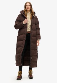 Brun lang pufferfrakke med hætte, quiltet design, lynlås foran og trykknapper; båret over en cremet sweater og sorte bukser med støvler.