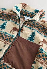 Kurtka z polaru w kremowym kolorze z wzorami gór i lasu w kolorach turkusowym, brązowym i ciemnozielonym; zawiera brązową kieszeń z tkaniny i zamek błyskawiczny.