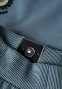 Tissu bleu clair avec un poignet côtelé. Comprend un bande élastique noir et un bouton circulaire argenté avec quatre trous.