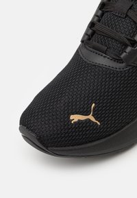 Crne atletske cipele s mrežastim gornjim dijelom, s teksturiranom površinom i zlatnim Puma logotipom sa strane. Glatka gumena potplata.