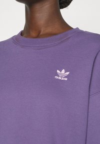 Fialová mikina s žebrovaným límcem a malým bílým logem Adidas na levé straně. Tkanina vypadá měkká a strukturovaná.