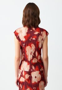Top plissettato floreale caratterizzato da colori rosso e crema con una superficie testurizzata, maniche a cappuccio e una vestibilità rilassata. I motivi includono grandi fiori.